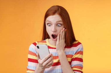 Omg ne oluyor. Portre şoke olmuş genç kızıl saçlı duyarlı kızıl saçlı kadın akıllı telefon görüntüsü dokunmatik yanak düşüşü şaşkına dönmüş turuncu arka planda duran şaşırmış telefon.