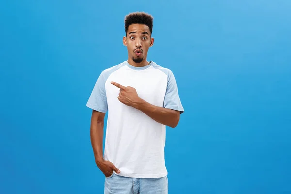 Impressionné curieux beau jeune homme avec barbe disant wow lever les sourcils dans l'émerveillement pointant vers la gauche étonné et excité debout ravi en t-shirt sur fond bleu — Photo