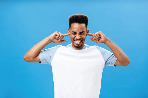 Irriterad och irriterad missnöjd ung afrikansk amerikansk kille i trendig t-shirt täcker öronen med pekfingrar rynkig näsa och blundar knyta tänderna från obehag höra högt bullriga ljud — Stockfoto