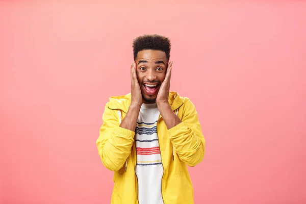 Non posso credere nella propria felicità. Sorpreso felice bello africano-americano studente maschio con acconciatura riccia e barba sorridente da eccitazione e brivido palme su guance sopra parete rosa — Foto Stock