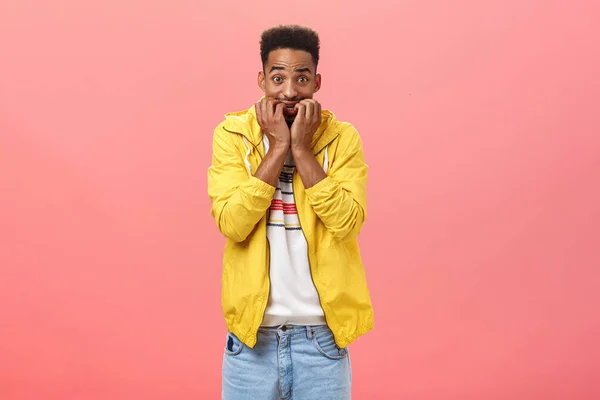 Indoor shot of worried young dark-skinned guy scared of resonsibilities starting panic biting fingernails raising hands and staring frightened at camera posing over pink background in yellow jacket — Stock Photo, Image