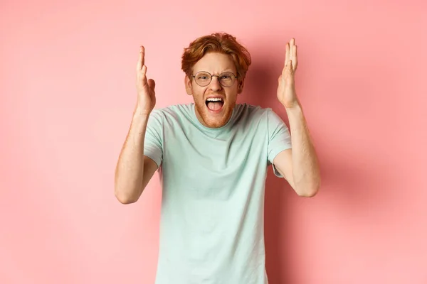 Boos roodharige man in bril schreeuwen, fronsen en schudden handen met gefrustreerd en verontwaardigd gezicht, staande over roze achtergrond — Stockfoto