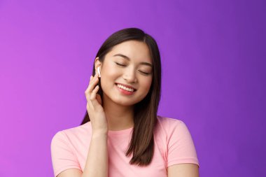 Şirin Asyalı kız müzik dinlemekten hoşlanıyor, kablosuz kulaklık takıyor, gözlerini kapatıyor rüya gibi gülümseyen tatmin oluyor, mükemmel ses çıkarıyor, kulaklığın sesini yükseltiyor, mor arka planda duruyor.