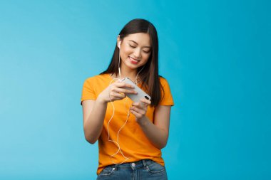 Hoş görünümlü şehir kızı tatilini yarış oyunu oynayarak geçiriyor, akıllı telefonu yatay pozisyonda tutuyor telefonu hareket ettiriyor, geniş bir coşkuyla gülümsüyor mavi arka plan.