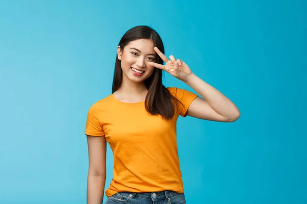 우정적 이고 명랑 한 동양인 여자 친구는 행복해 보이고, 평화의 승리의 표시를 보이고, 긍정적 인 미소를 짓고, 운 이 좋은 여름 화창 한 날을 즐기고, 푸른 배경에 푸른 티셔츠를 입고 서 있다 — 스톡 사진