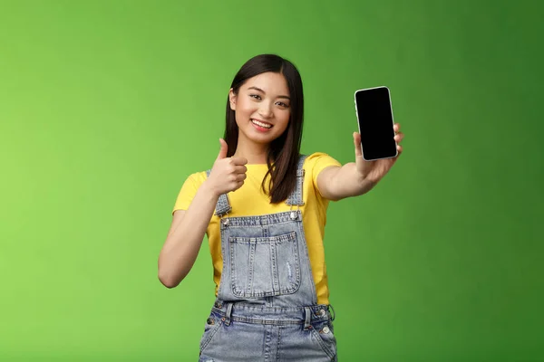Glada söt asiatisk kvinna att bedöma bra smartphone spel, visa tummen upp som tecken, förlänga arm håll telefonskärm, presentera awesome app, främja ansökan, stå grön bakgrund — Stockfoto