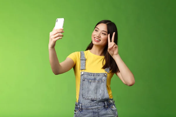 Tierna mujer morena asiática tierna, mantenga el teléfono inteligente, posando selfie, mira la pantalla del teléfono, fotografiando, mostrar señal de paz victoria, alegre charla amigo en el extranjero, de pie fondo verde — Foto de Stock