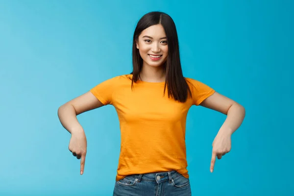 Fröhlich selbstbewusste asiatische Mädchen mit dunklem Haarschnitt tragen gelbes Sommer-T-Shirt, führen Promo ein, zeigen die Hände nach unten, zeigen unten Werbung lächelnd erfreut, teilen guten Link blauen Hintergrund — Stockfoto