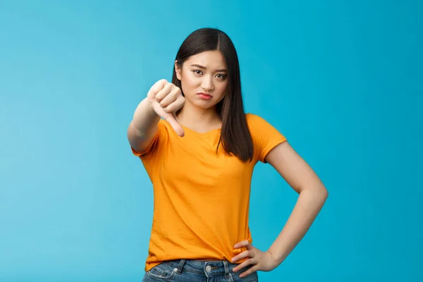 Csalódott és elégedetlen aranyos ázsiai lány grimaszoló, mutató hüvelykujj-down nem, nem tetszik jel, homlokráncolás, remegő idegenkedés és vonakodás, állni elégedetlen kék háttér — Stock Fotó