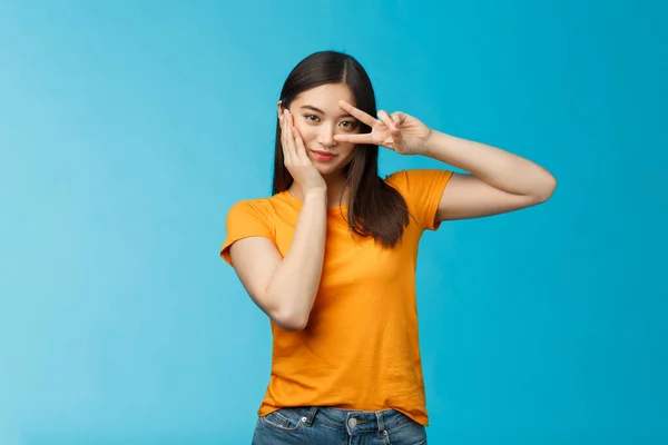 Descarado elegante de buen aspecto asiático asertivo coreano chica mostrar paz victoria signo en el ojo, toque mejilla mirada empoderado y motivado, de pie descarado coqueta azul fondo, desgaste amarillo camiseta —  Fotos de Stock