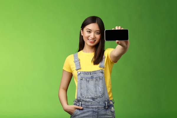 Attraktiv glad asiatisk brunett kvinna införa smartphone spel, hålla telefonen horisontellt, visa mobiltelefon display, ler glad, ge rekommendation vilken app nedladdning, grön bakgrund — Stockfoto