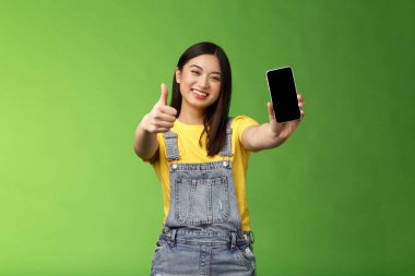 Neşeli, tatlı Asyalı esmer kız harika bir akıllı telefon oyunu, telefon ekranını göster, işaret ver, geniş gülümse, harika online uygulamayı tanıt, yeşil arka plan