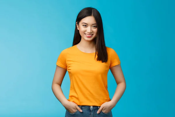 Självsäker övertygande leende asiatisk kvinna stå blå bakgrund look kamera, hålla händer jeans fickor uttrycka självsäker vibb, njuta av att se positiva resultat hudvård förfaranden, bära gul t-shirt — Stockfoto