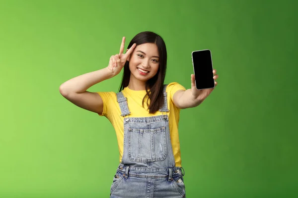 Söt bekymmerslös asiatisk brunett visar ansökan på smartphone skärmen, göra seger fred tecken, ler glatt, skryta sociala medier popularitet, anhängare belopp, stå grön bakgrund — Stockfoto