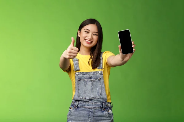 Glada nöjd söt asiatisk brunett flicka nöjd awesome smartphone spel, visa telefonskärm, ge tummen upp som tecken, ler brett, införa awesome online-program, grön bakgrund — Stockfoto