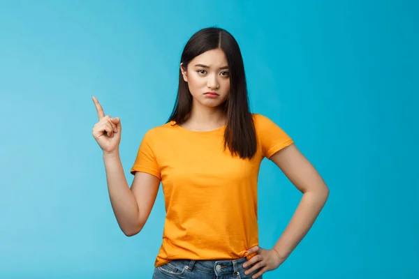 Муді засмучена азіатська дівчина, засмучений виглядає ображеним і незадоволеним, вказуючи верхній лівий кут, кидаючи розчарований, стоячи засмучений синій фон одягнути жовту футболку — стокове фото