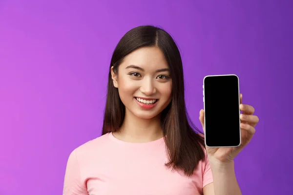 Close-up attractive pleasant female shop assistant present new phone app, hold smartphone show telephone screen, introduce application on display, stand purple background delighted