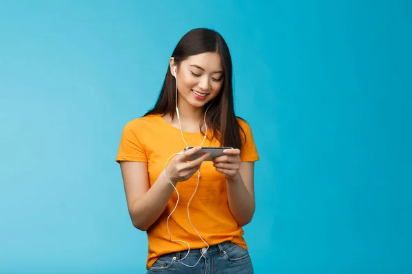 Feliz entretenido asiática linda chica morena, usar auriculares no molestó a la gente en la cafetería mientras jugaba juego de teléfono inteligente favorito, mantener el teléfono horizontal, sonrisa mirada entusiasta pantalla del teléfono celular — Foto de Stock