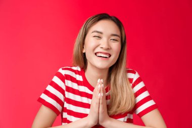 Yakın plan neşeli kaygısız sevimli Asyalı kız el ele tutuşuyor dua ediyor, arkadaşına teşekkür ediyor gülümseyerek gülüyor, mutlu bir ruh hali içinde iyilik istiyor, yardım dileniyor kırmızı arka plan için yalvarıyor