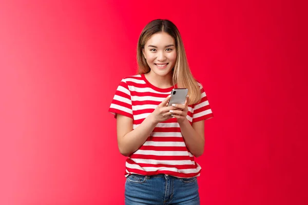 Glada ganska asiatisk blond flicka fnissande leende toothy, känna sig glad, hålla smartphone läsa vän roligt meddelande, stå röd bakgrund nonchalant, svepa dejting app, handla online butik bekymmersfri — Stockfoto