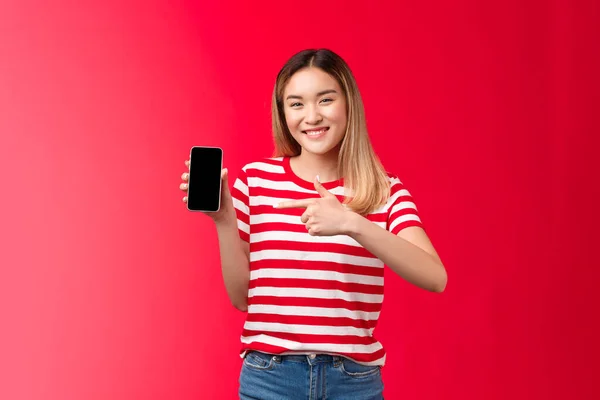 Molto soddisfatto allegro carino ragazza bionda asiatica sorridente ampiamente mostra display smartphone, schermo del telefono a punta contento introdurre impressionante nuova applicazione, raccomandare l'uso dell'applicazione, sfondo rosso — Foto Stock