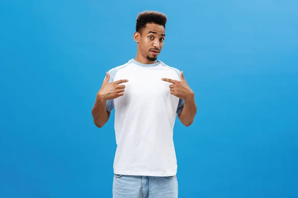 Le gars se sent surpris d'être choisi par le manager. Portrait d'un beau jeune homme afro-américain stupéfait à la barbe et à la coupe de cheveux bouclée se pointant vers lui avec un regard interrogé sur le mur bleu — Photo