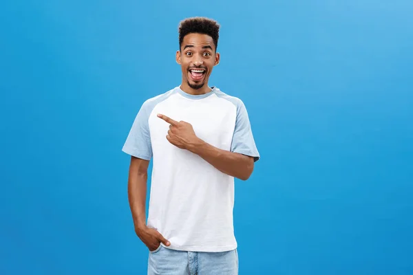 Enthusiastic pleased and charmed dark-skinned guy with beard and afro hairstyle standing joyful over blue background smiling joyfully pointing at upper left corner holding hand in pocket — Stock Photo, Image