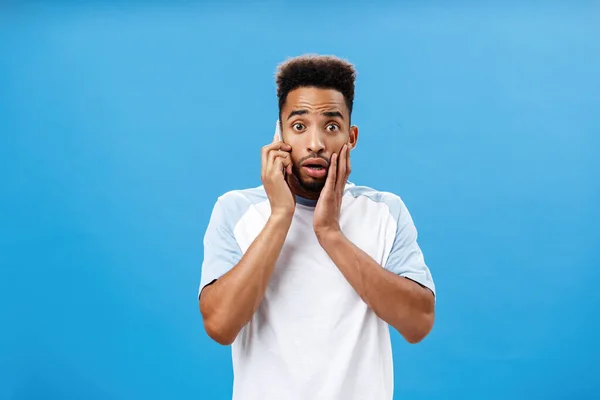 Homem pegando telefone ouvir notícias terríveis sentindo-se chocado e preocupado segurando smartphone perto da boca de abertura do ouvido, franzindo a testa e tocando o rosto do choque preocupante para a saúde do amigo sobre a parede azul — Fotografia de Stock