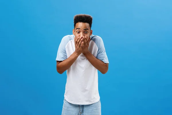 Shocked young dark-skinned male student witnessing murder screaming and starting panic covering mouth with both palms to calm down raising eyebrows staring with fear and fright over blue wall — Stock Photo, Image