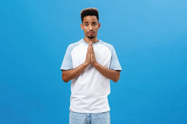 Please help me beg you. Charming cute and handsome african american boyfriend with afro curly haircut and beard holding hands in pray frowning making silly face while asking favor over blue wall — Stock Photo, Image