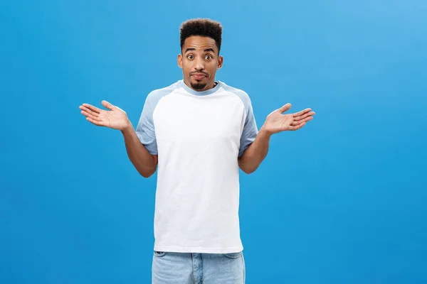 Studio shot of unaware clueless cute african american guy with beard and afro hairstyle shrugging with palms spread aside pursing lips and stooping while being unsure and uncertain over blue wall — Stock Photo, Image