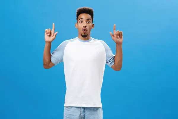 Impressionado emocionado bonito namorado afro-americano em casual t-shirt lábios dobráveis em uau som sendo surpreendido e surpreso com o produto legal apontando para cima com as mãos levantadas sobre a parede azul — Fotografia de Stock