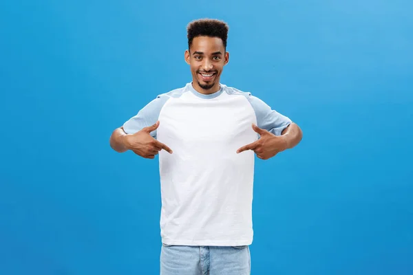 Agradável e encantado homem de boa aparência mostrando t-shirt legal apontando para si mesmo com expressão satisfeita orgulhoso em pé sobre fundo azul feliz e amigável, orgulhosamente contando sobre seus objetivos — Fotografia de Stock