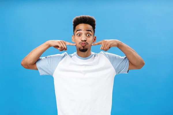 Waist-up shot of playful happy nice african american guy with tattoo on palm holding breath pouting joyfully and poking at cheeks with index fingers popping eyes while having fun over blue background — Stock Photo, Image