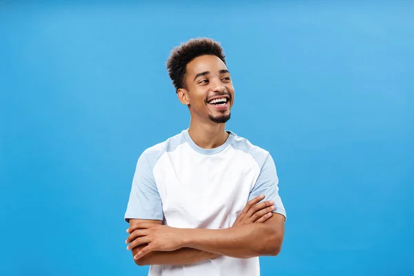 Proud and carefree cool african american male student in trendy t-shirt holding hands crossed on chest in self-assured carefree gesture turning right laughing pleased, having fun over blue wall — Stock Photo, Image