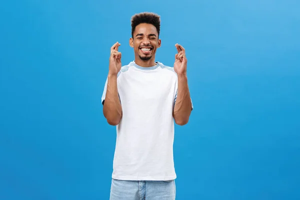 Putting all effort in pray to make dream come true. Portrait of hopeful intense and optimistic dark-skinned guy with afro haircut smiling closing eyes crossing fingers for good luck over blue wall — Stock Photo, Image