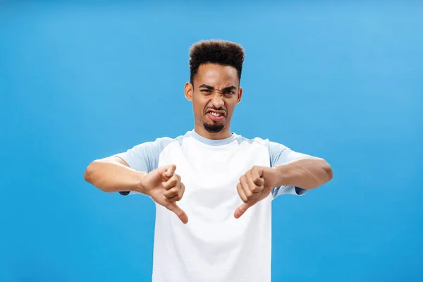 Disgusted dissatisfied dark-skinned young guy with beard and afro hairstyle frowning wrinkling nose and showing thumbs down with dislike expressing disapproval, giving negative feedback — Stock Photo, Image