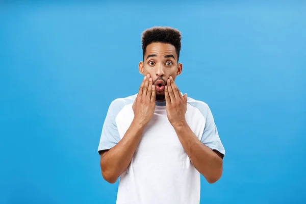 Attractive young dark-skinned guy likes hearing fresh rumors learning new hot details gasping opening mouth holding hands on face staring impressed and astonished at camera over blue background — Stock Photo, Image