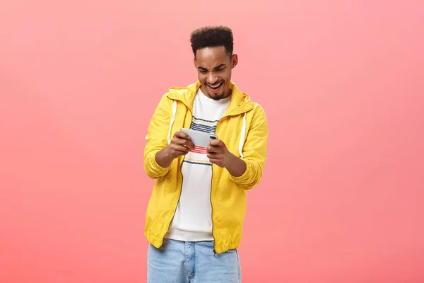 Enthusiastischer Typ, der Spaß beim Spielen eines fantastischen Smartphone-Spiels hat, das Handy in beiden Händen hält und mit Leidenschaft und Nervenkitzel auf den Bildschirm starrt, verbringt seine Freizeit im Internet vor rosa Hintergrund — Stockfoto