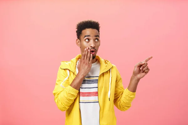 Shocked thrilled african american male with beard and afro haircut saying wow with opened mouth and palm on lip pointing and gazing at upper right corner intrigued over pink background — Stock Photo, Image