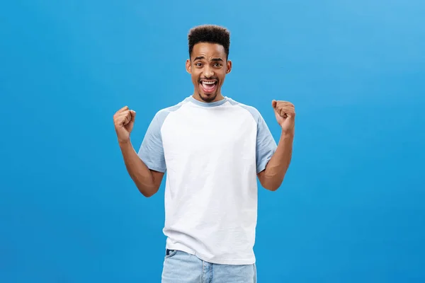 Ondersteunende en juichen Afrikaans-Amerikaanse jonge sporter in trendy t-shirt hef vuisten in de overwinning en triomf gebaar schreeuwen ja met vreugde en tevredenheid vieren, het ontvangen van positief resultaat — Stockfoto