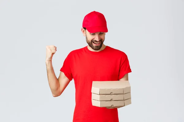 Livsmedelsleverans, karantän, stanna hemma och beställ online koncept. Glad, upphetsad skäggig kurir i röd t-shirt och mössa firar, glädje, tittar på pizza order, grå bakgrund — Stockfoto