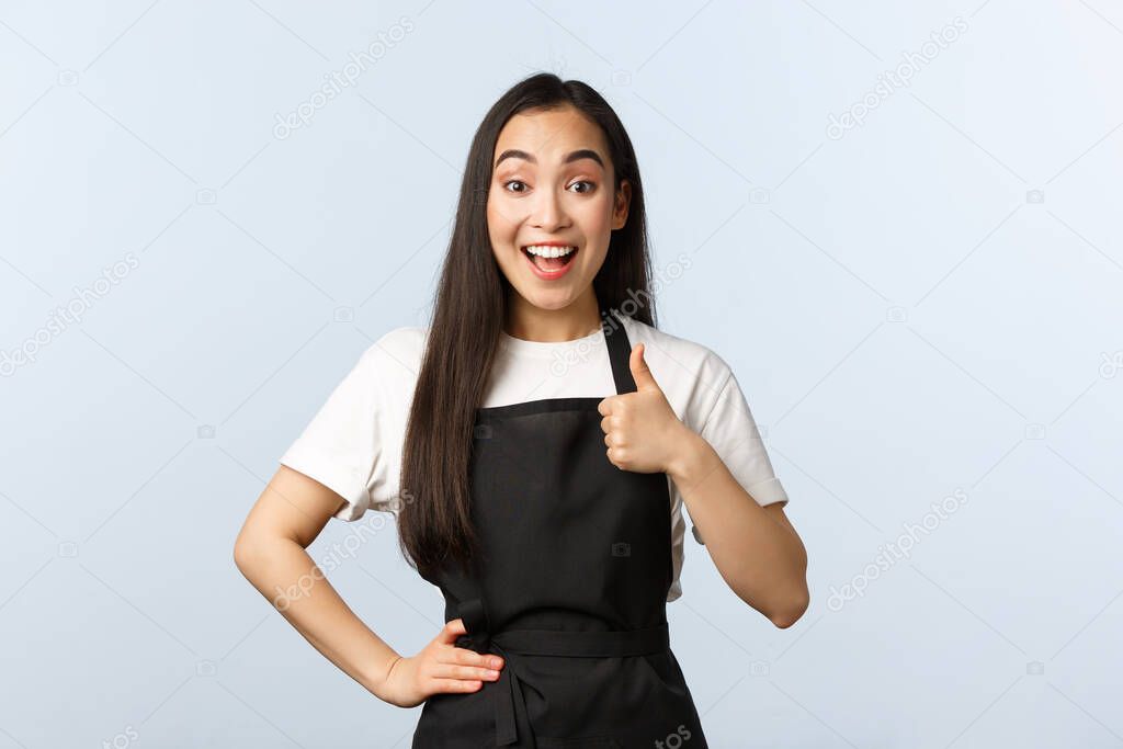 Coffee shop, small business and startup concept. Cheerful young pretty female cafe staff in black apron show support or like gesture. Asian waitress make thumb-up and smiling satisfied