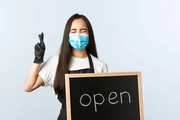 Småföretag, covid-19 pandemi, virus och anställda koncept. Spännande och optimistisk asiatisk barista, kvinnlig anställd i medicinsk mask, visar öppen skylt och ber lycka till, långfingrar — Stockfoto