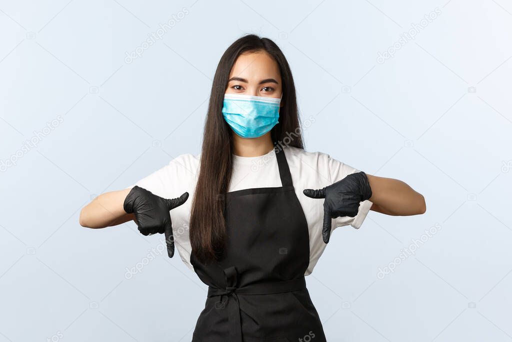 Covid-19 pandemic, social distancing, small business and preventing virus concept. Young pleasant asian barista, female coffee shop owner inviting to cafe, pointing down, wear medical mask and gloves