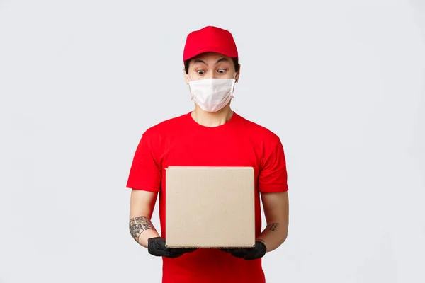 Здивований азіатський хлопець доставки дивиться на посилку з дивним шумом. Кур'єр одягнений у червону шапку та футболку з захисними рукавичками, медичною маскою. Надання клієнту свого замовлення ковадла-19 самокарантину — стокове фото