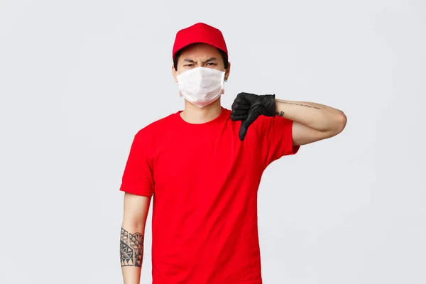 Une autre livraison horrible. Réticent, déçu livreur en casquette rouge, t-shirt, portant des gants de protection et un masque médical, grimaçant aversion et désaccord, montrer pouce vers le bas mauvaise opinion négative — Photo