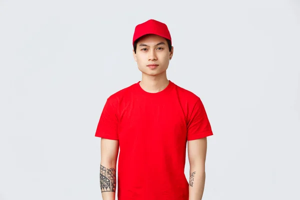 Expressz szállítási, szállítási és logisztikai koncepció. Fiatal jóképű ázsiai futár piros sapkában és pólóban, figyelő kamera, várja az ügyfél megrendelés szállított vállalati tárolás — Stock Fotó