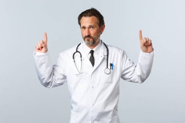 Covid-19, koronavirüs salgını, sağlık çalışanları ve pandemik konsept. Beyaz önlüklü kuşkucu yetişkin erkek doktor kamerayı işaret ediyor ve endişeli görünüyor. Şüpheleri var.