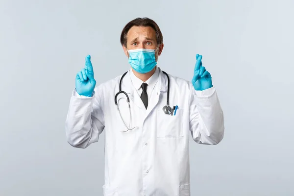 Covid-19, πρόληψη του ιού, εργαζόμενοι στον τομέα της υγείας και την έννοια του εμβολιασμού. Προβληματικός αισιόδοξος γιατρός με ιατρική μάσκα και γάντια, σταυρωμένα δάχτυλα καλή τύχη, προσευχή ή ευχή, λευκό φόντο — Φωτογραφία Αρχείου